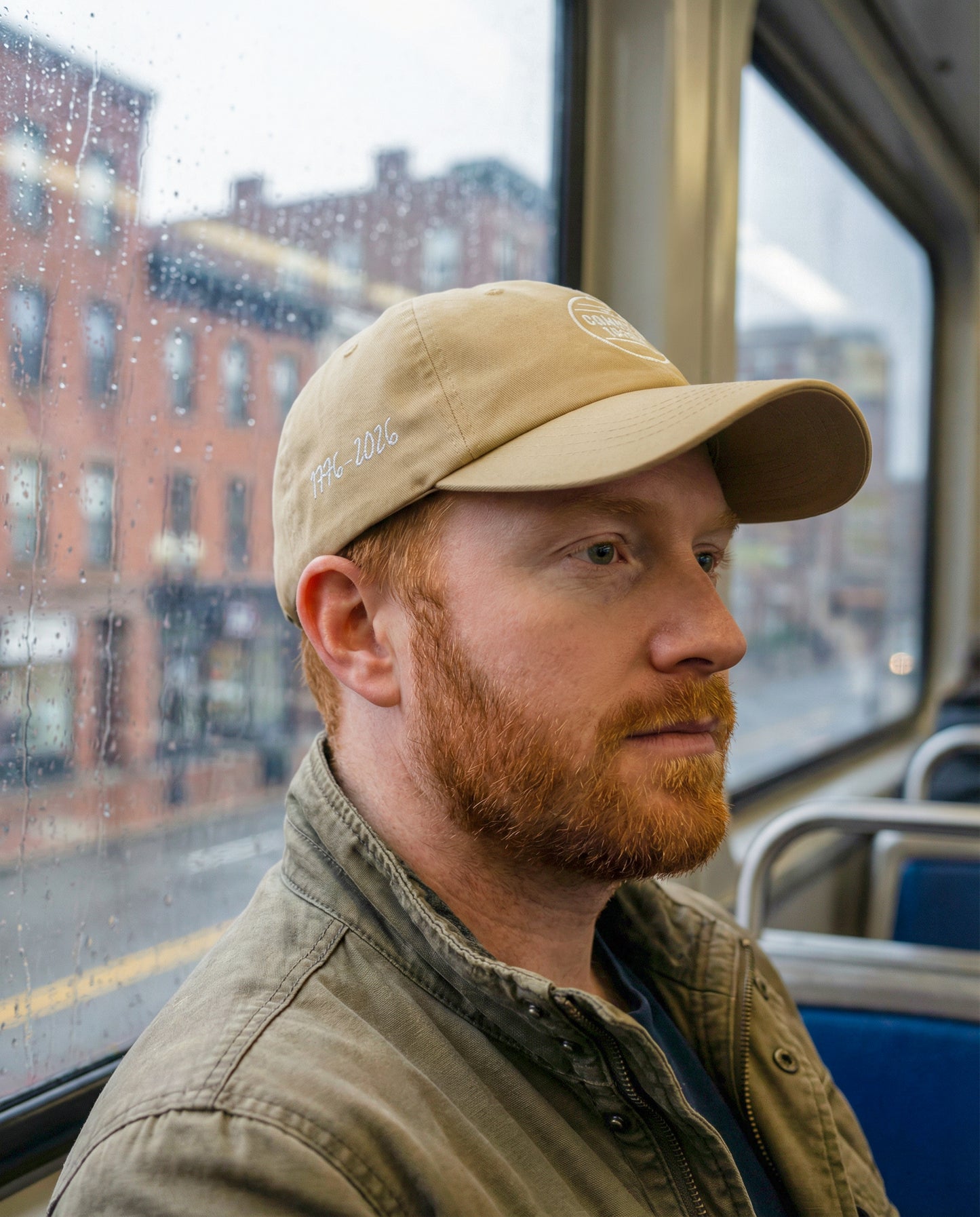 Man with a beard wearing a beige cap and jacket on a bus, looking out the window.