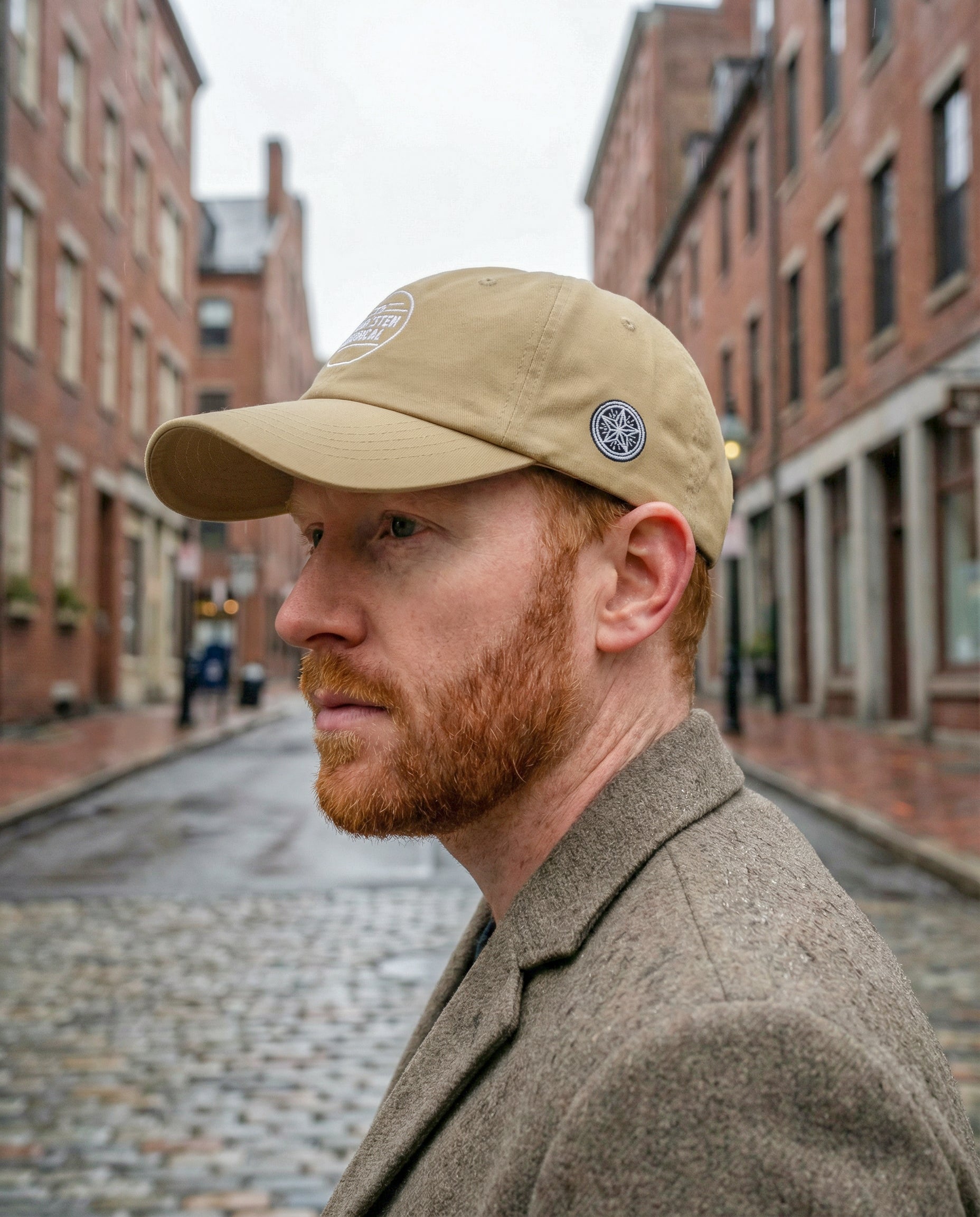 Man wearing a beige cap and coat on a street with brick buildings.