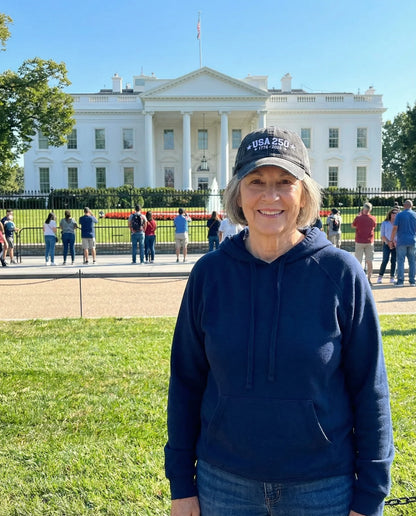Person standing in front of the White House wearing a blue hoodie and a cap with 'USA 250' text.