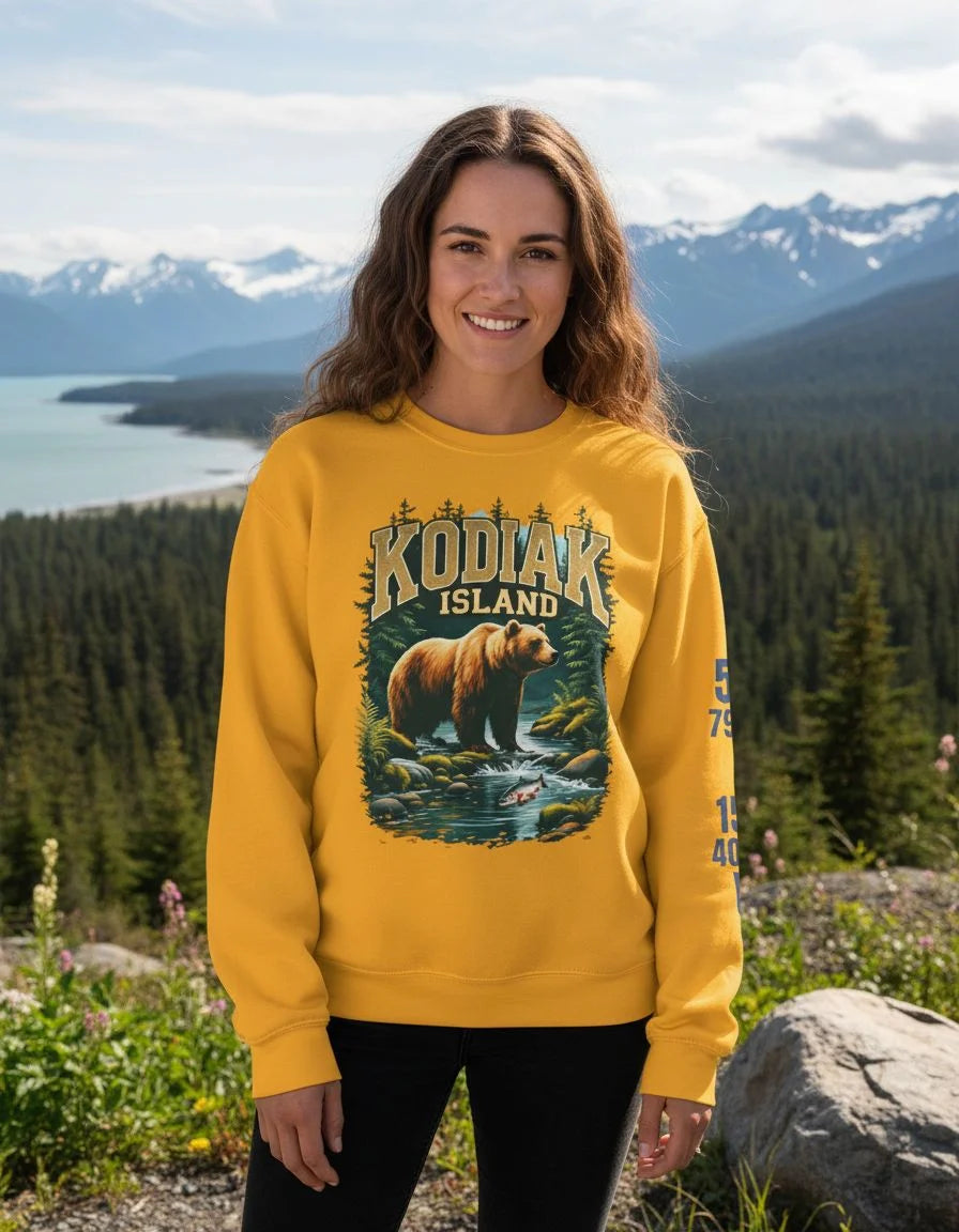 Woman wearing a yellow sweatshirt with a bear graphic and 'Kodiak Island' text, standing in front of a scenic mountain landscape.