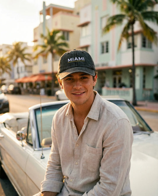 Man wearing a 'Miami' cap sitting on a classic car with palm trees and buildings in the background