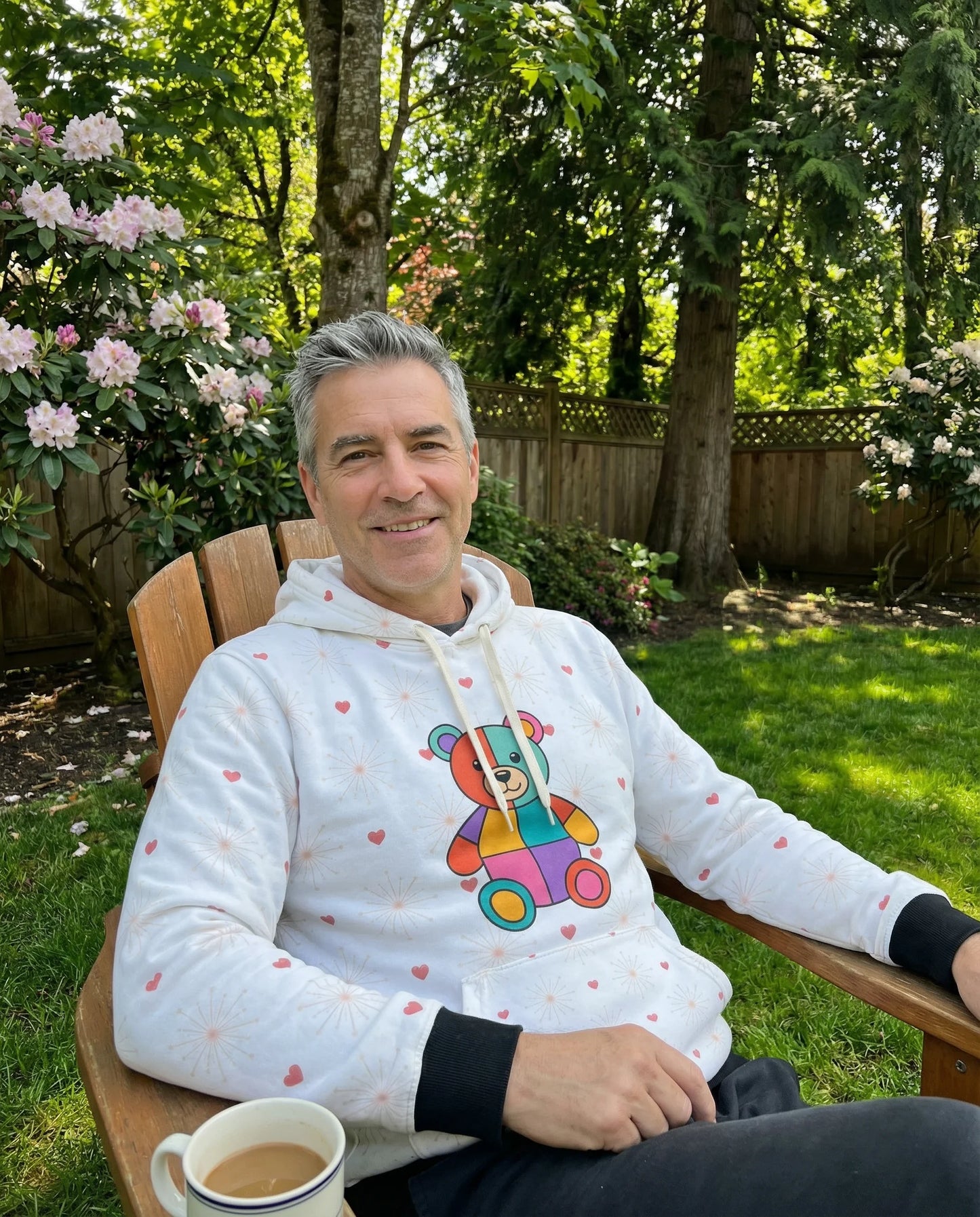Man sitting outdoors in a garden wearing a white hoodie with a colorful graphic, holding a cup.