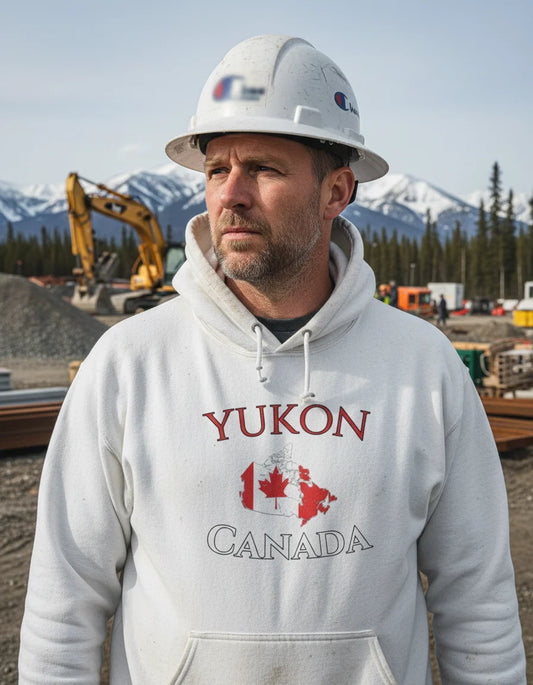 Man wearing a 'Yukon Canada' hoodie with a hard hat, standing in a construction site with mountains in the background.